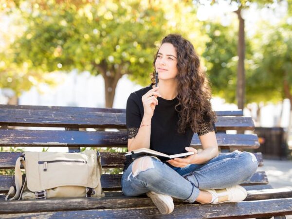 Cute Young Woman Sitting Outside In A Park Bench And Thinking While Writing In A Journal